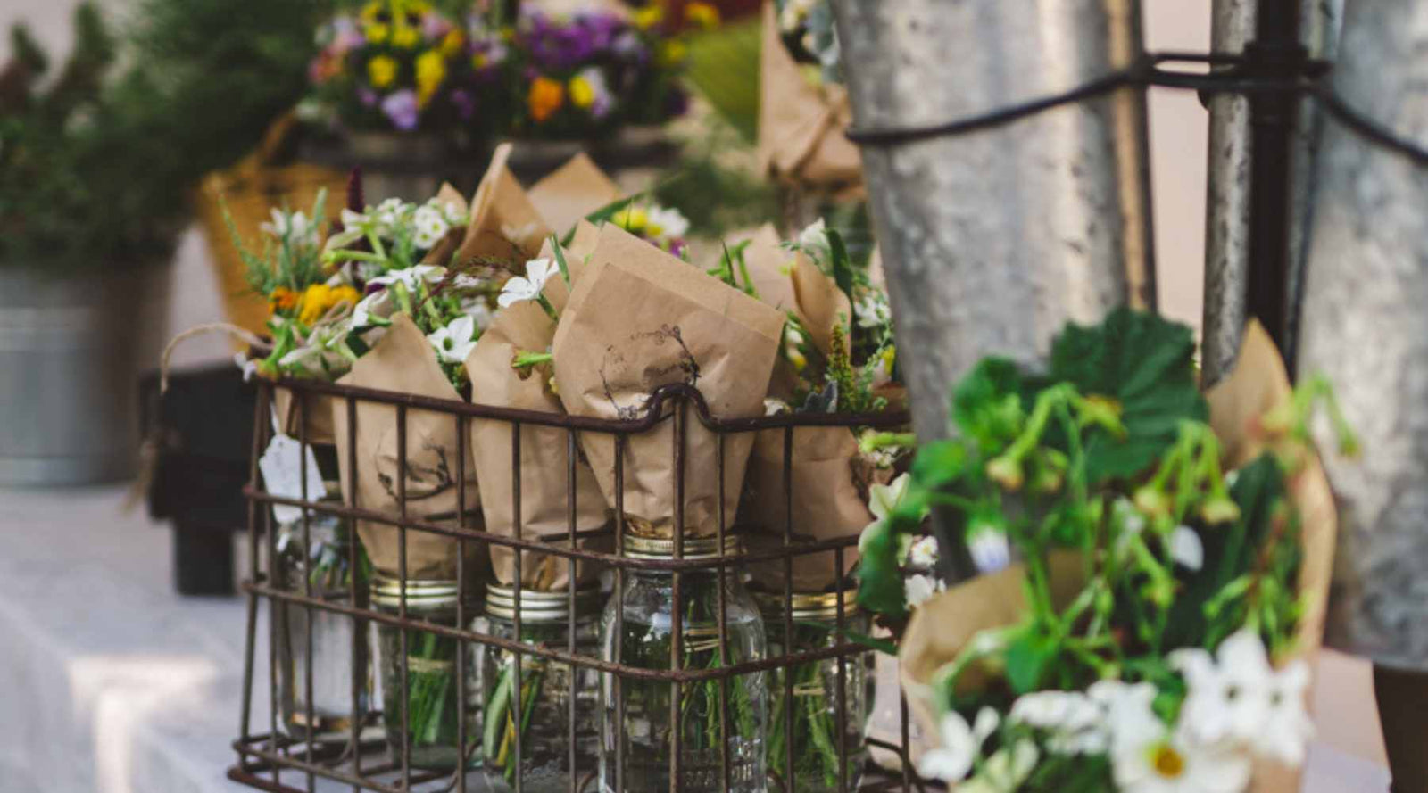 Bouquets of flowers wrapped in brown paper and placed in mason jars. The jarred bouquets are inside of a wire basket sitting on a curb.
