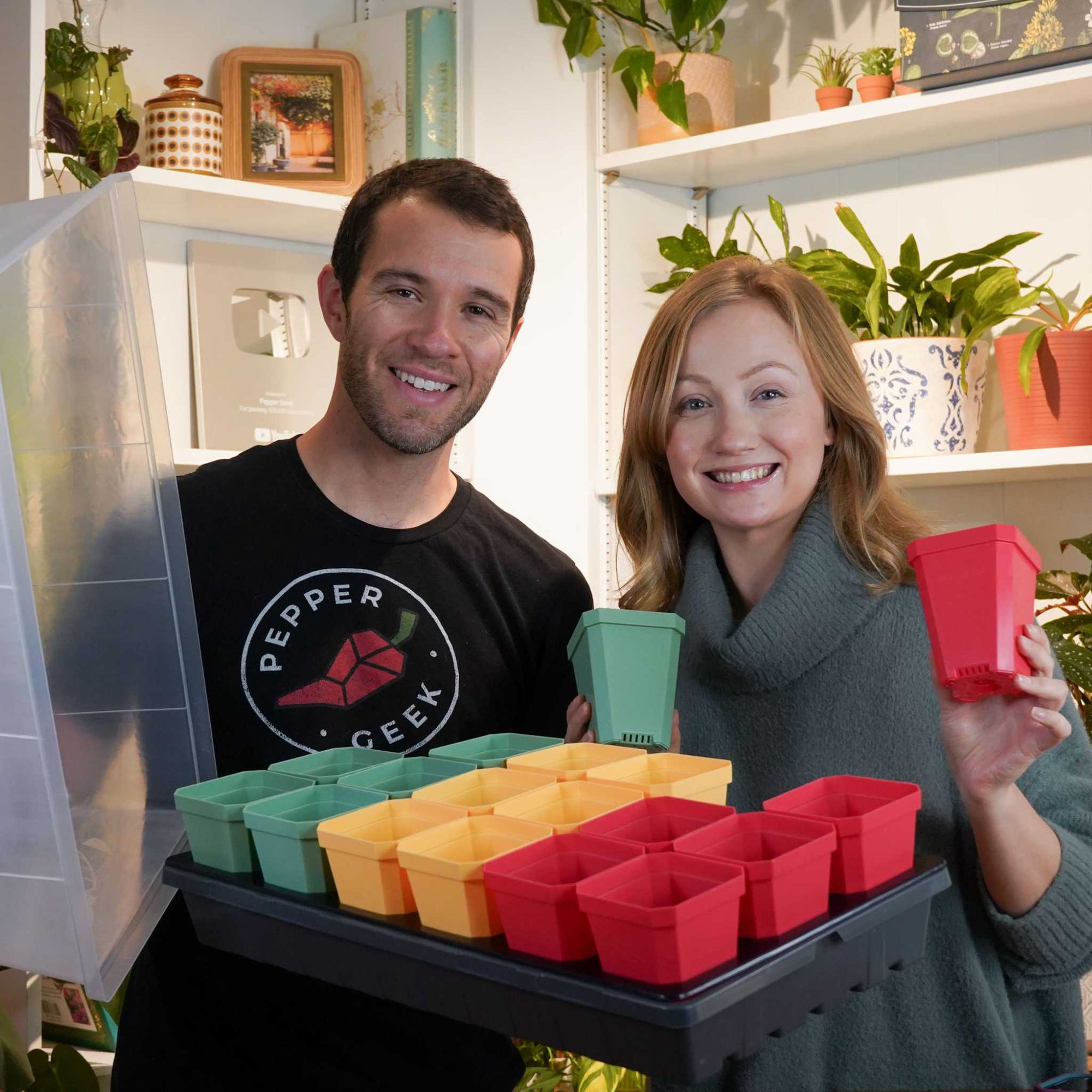 Woman holding a red and a green 3.3 inch pot. She is standing next to a man wearing a Pepper Geek shirt and holding a grey 1020 full of 3 inch pots.