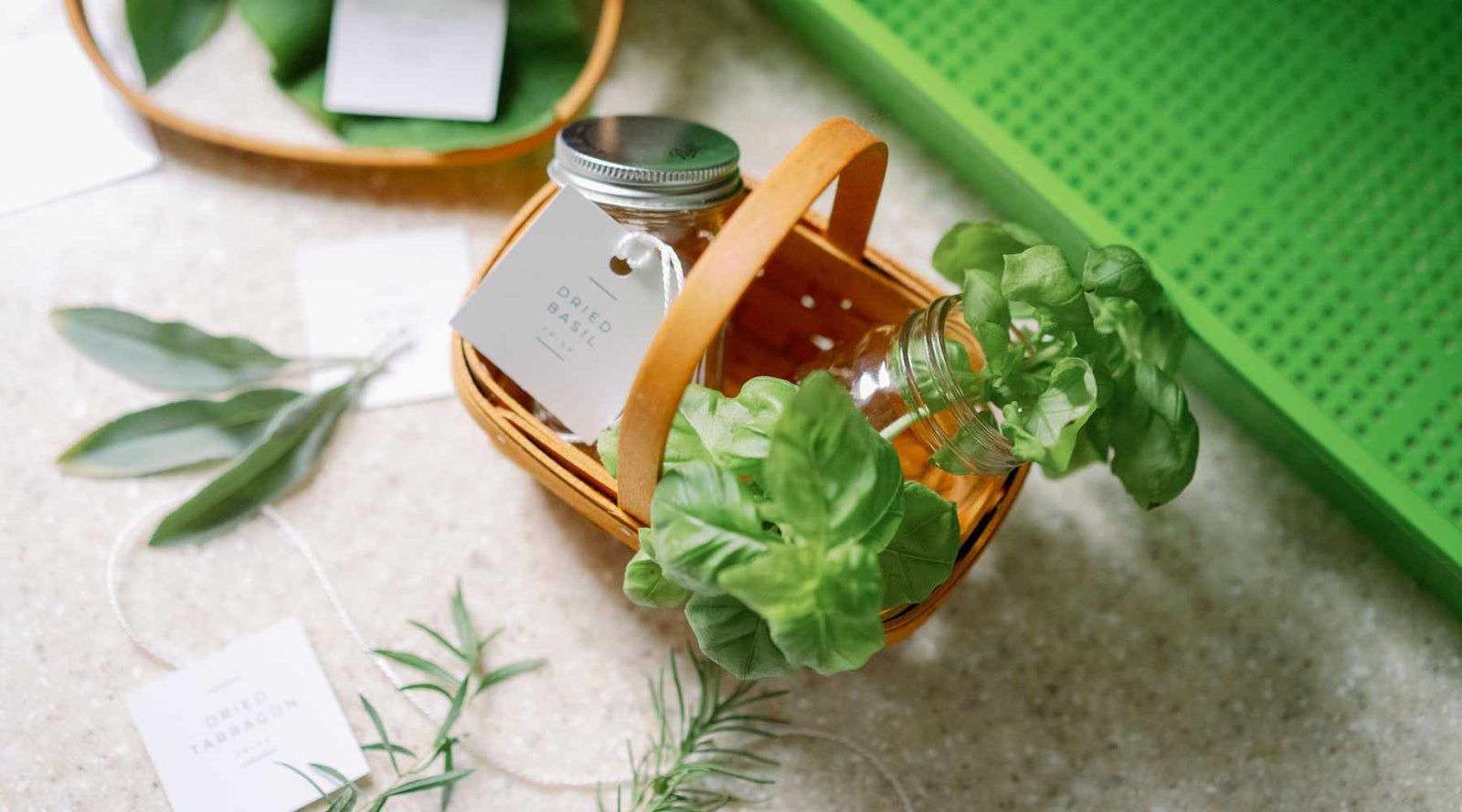 A small basket holding bunches of basil with sprigs of rosemary, sage and a green mesh tray in the background. 