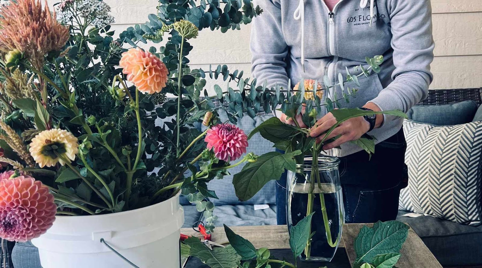 A white bucket full of dahlias and foliage in the foreground. a person is visible in the background arranging flowers in a tall glass vase. 