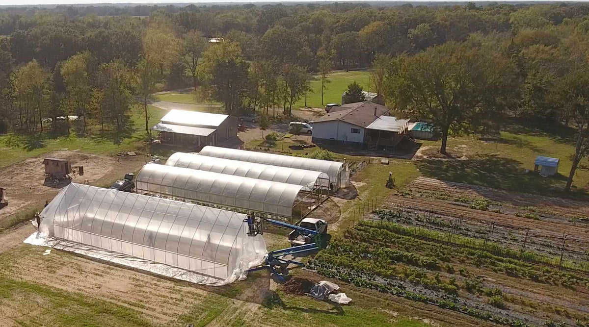 Aerial view of a farm with four high tunnels and rows of market garden crops. The closest hoop frame has a scissor lift next to it and is in the process of being covered with greenhouse plastic.   