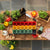 Overhead view of a wooden planting table with red, green, and yellow 3 inch pots in a dark grey 1020 tray with insert holder. Also on the table are young plants, peppers, watering can, and pepper seeds.